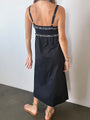 Cropped chin down back view of woman in a black midi length tank dress with lace bodice and white embroidered ruffle trim details standing against plain background. 