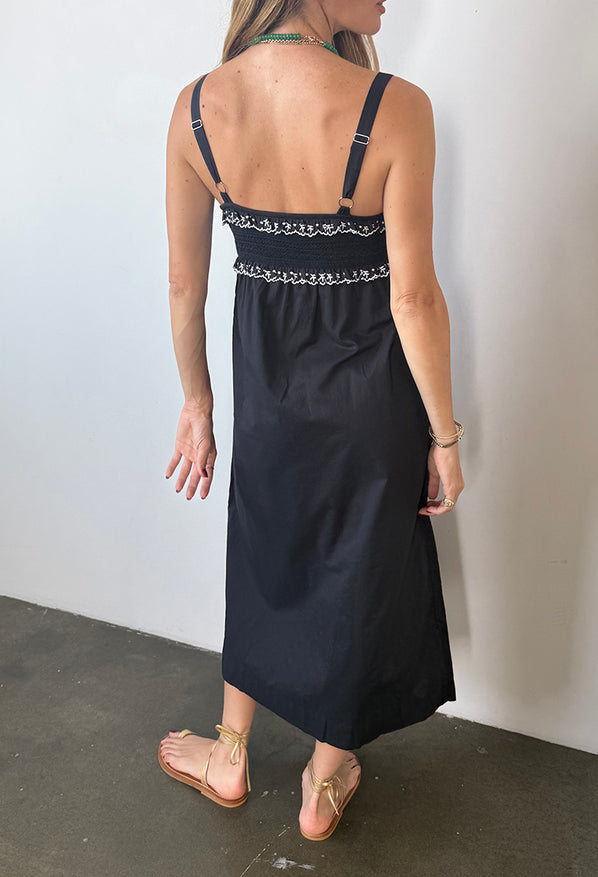 Cropped chin down back view of woman in a black midi length tank dress with lace bodice and white embroidered ruffle trim details standing against plain background. 