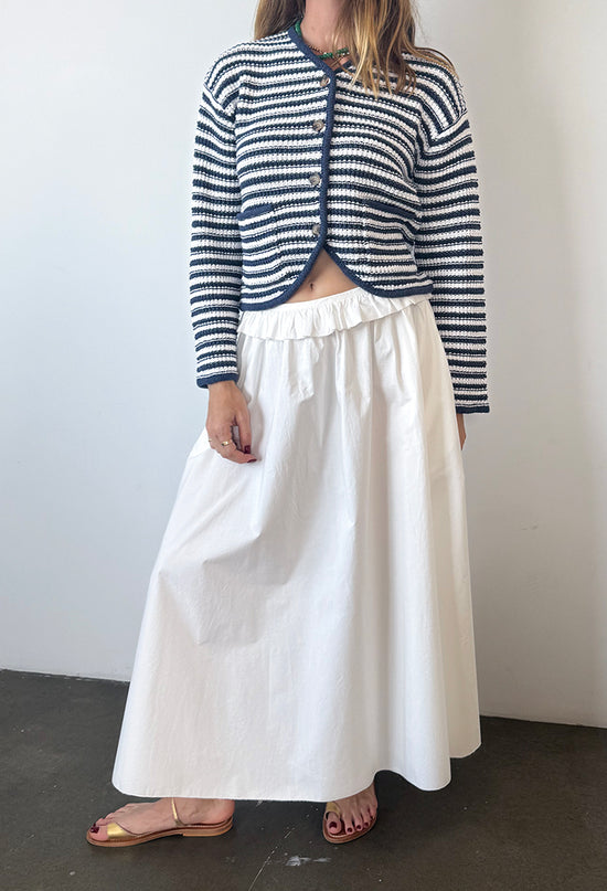 Cropped neck down of woman wearing a striped cardigan and long white skirt with ruffles along the waistline against a plain background