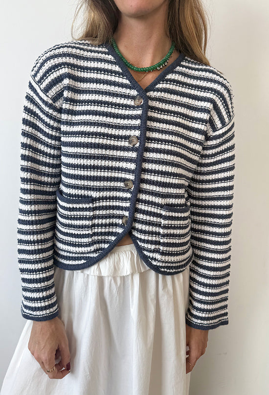 Cropped chin to thigh of woman wearing a striped white and navy knit cardigan with buttons against a white background. 