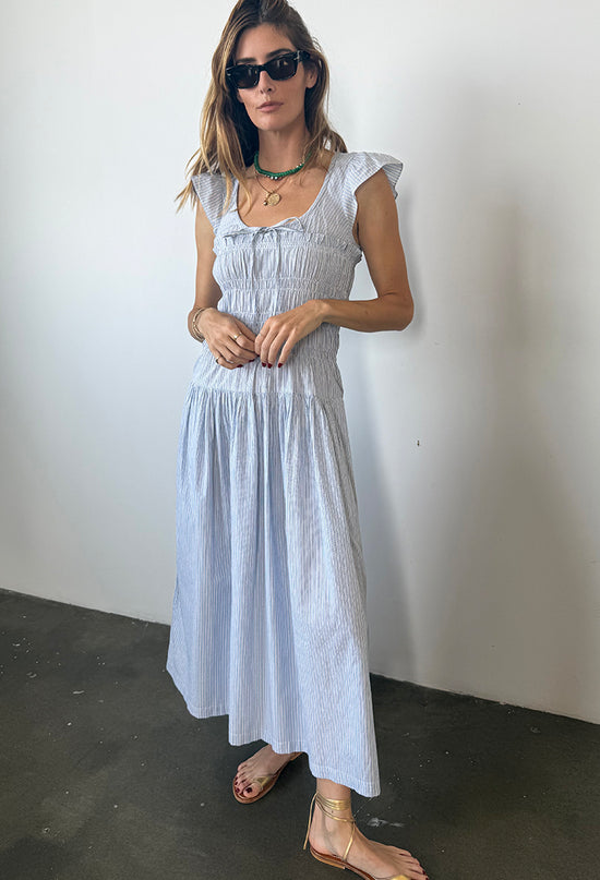 Woman wearing a light blue pinstriped short-sleeved midi dress with a scoop neckline with a tie and smocking around the bust to the waist against a white wall