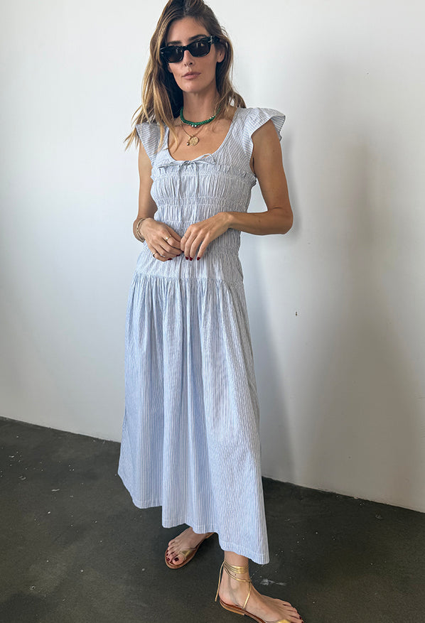 Woman wearing a light blue pinstriped short-sleeved midi dress with a scoop neckline with a tie and smocking around the bust to the waist against a white wall