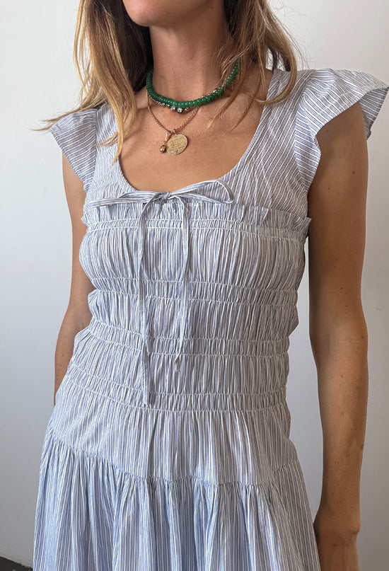 Cropped chin to waist of woman wearing a light blue pinstriped short-sleeved midi dress with a scoop neckline with a tie and smocking around the bust to the waist against a white wall