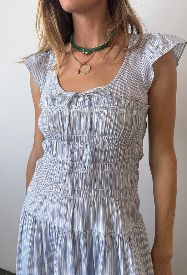 Cropped chin to waist of woman wearing a light blue pinstriped short-sleeved midi dress with a scoop neckline with a tie and smocking around the bust to the waist against a white wall