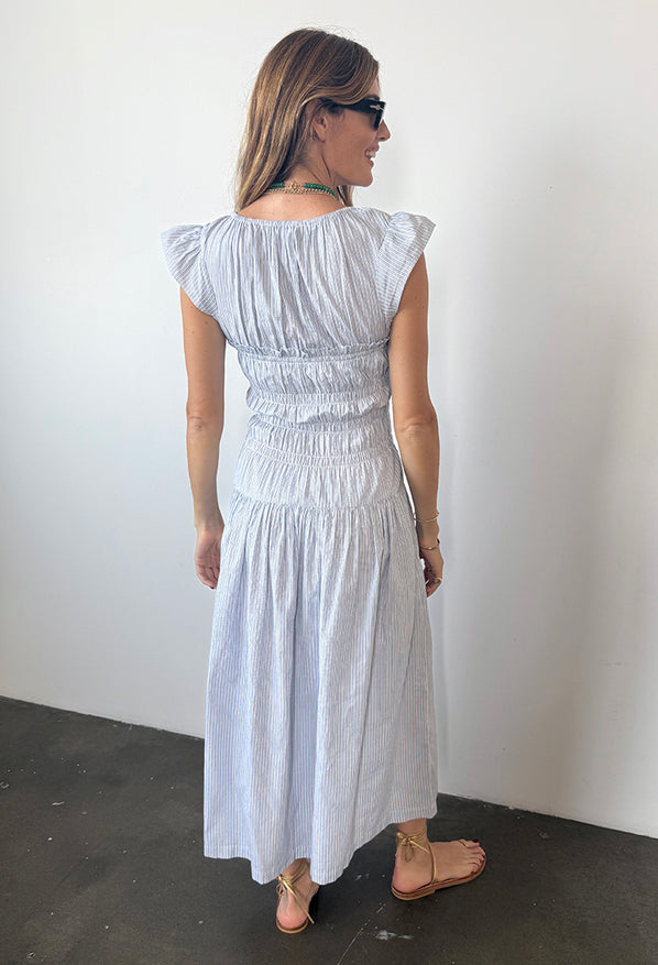 Back view of woman wearing a light blue pinstriped short-sleeved midi dress with smocking around the bust to the waist against a white wall