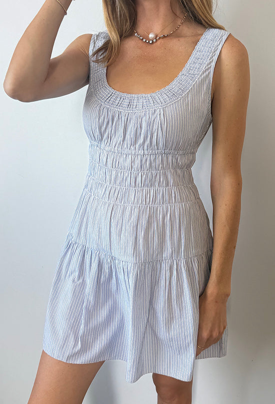 Cropped neck to knee of woman in a light blue pin-striped tank style mini dress with a smocked bodice against a plain background