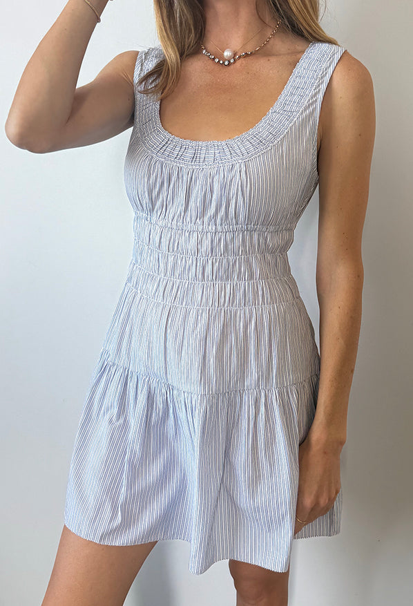 Cropped neck to knee of woman in a light blue pin-striped tank style mini dress with a smocked bodice against a plain background