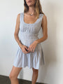 Image cropped nose to ankles of woman in a light blue pin-striped tank style mini dress with a smocked bodice standing against a white wall