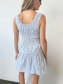 Image cropped head to knee of woman in a light blue pin-striped tank style mini dress with a smocked bodice standing against a white wall