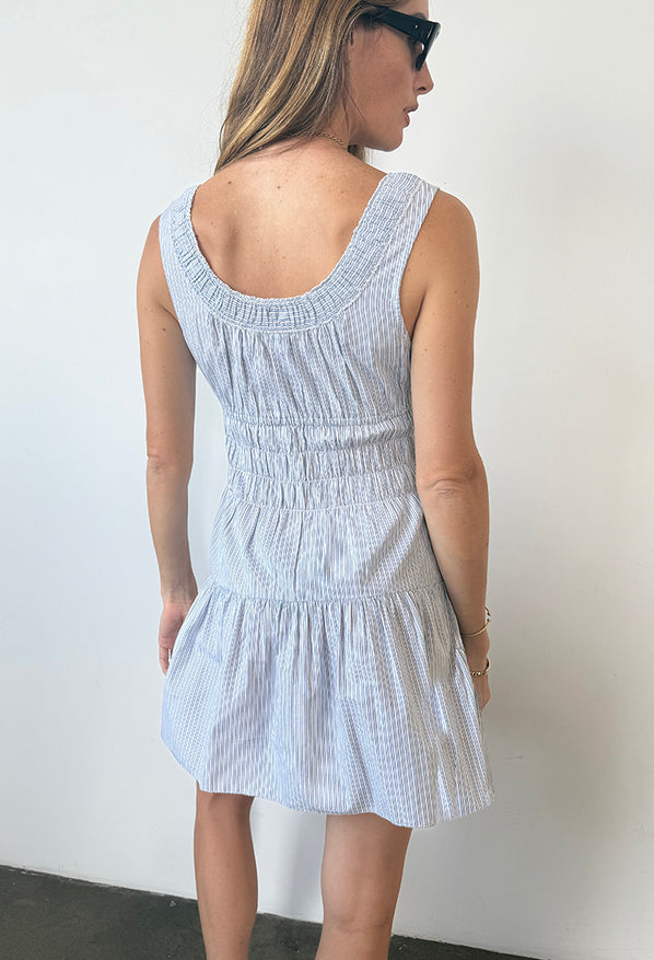 Image cropped head to knee of woman in a light blue pin-striped tank style mini dress with a smocked bodice standing against a white wall