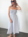 Woman in a light blue pin-striped midi length dress with spaghetti straps, smocked bodice and ruffled neckline and waistline against a white background