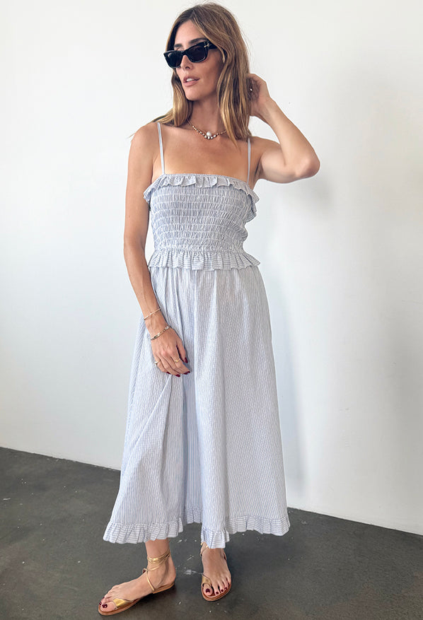 Woman in a light blue pin-striped midi length dress with spaghetti straps, smocked bodice and ruffled neckline and waistline against a white background