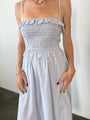 Image cropped shoulders to knee of woman in a light blue pin-striped midi length dress with spaghetti straps, smocked bodice and ruffled neckline and waistline against a white background