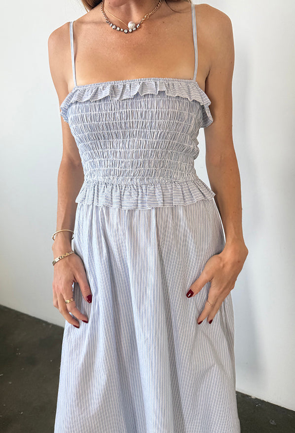 Image cropped shoulders to knee of woman in a light blue pin-striped midi length dress with spaghetti straps, smocked bodice and ruffled neckline and waistline against a white background
