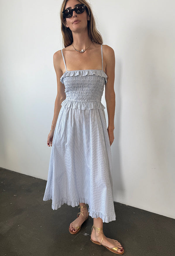 Woman in a light blue pin-striped midi length dress with spaghetti straps, smocked bodice and ruffled neckline and waistline against a white background