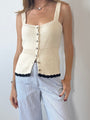 Cropped chin to thigh of woman wearing a cream knitted button up tank top with black hem details paired with light blue pin-striped pants against a plain background. 