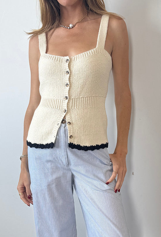 Cropped chin to thigh of woman wearing a cream knitted button up tank top with black hem details paired with light blue pin-striped pants against a plain background. 