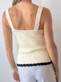 Cropped chin to thigh of woman wearing a cream knitted button up tank top with black hem details paired with light blue pin-striped pants against a plain background. 