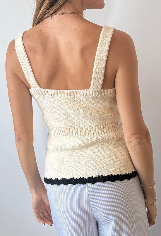 Cropped chin to thigh of woman wearing a cream knitted button up tank top with black hem details paired with light blue pin-striped pants against a plain background. 