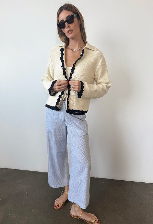Woman wearing a beige knit sweater / jacket with black trim and light blue pin-striped wide leg pants against a white wall.