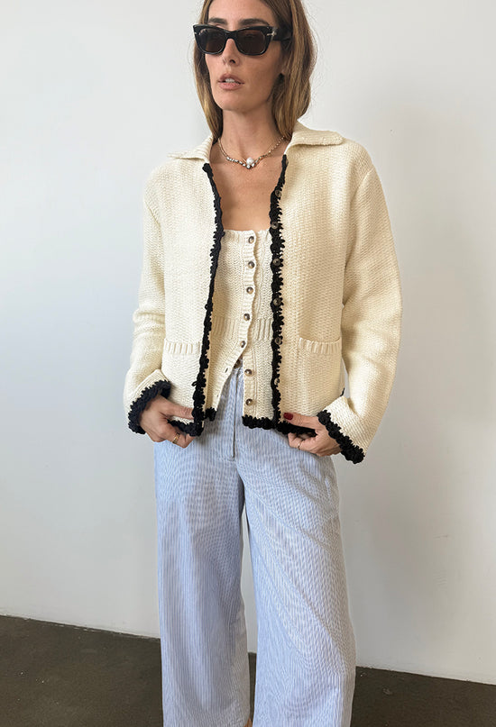 Cropped forehead to shin of woman in ivory knit jacket with black trim detailing along the sleeves, hem, and center seam and light blue pin-striped pants against a white background