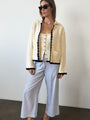 Woman in ivory knit jacket with black trim detailing along the sleeves, hem, and center seam and light blue pin-striped pants against a white background