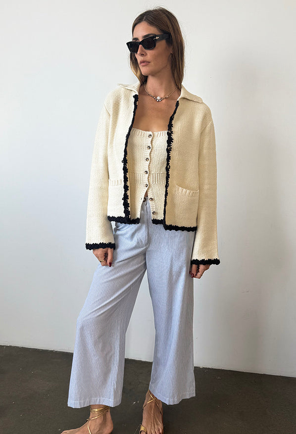 Woman in ivory knit jacket with black trim detailing along the sleeves, hem, and center seam and light blue pin-striped pants against a white background