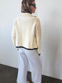 Woman from behind view in ivory knit jacket with black trim detailing along the sleeves, hem, and center seam and light blue pin-striped pants against a white background