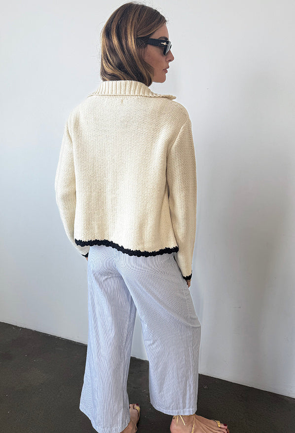 Woman from behind view in ivory knit jacket with black trim detailing along the sleeves, hem, and center seam and light blue pin-striped pants against a white background