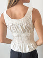 Cropped nose to waist of woman wearing a white thick strapped tank top with a shirred scoop neckline and tiered rows of smocking around the waist against a white background. 