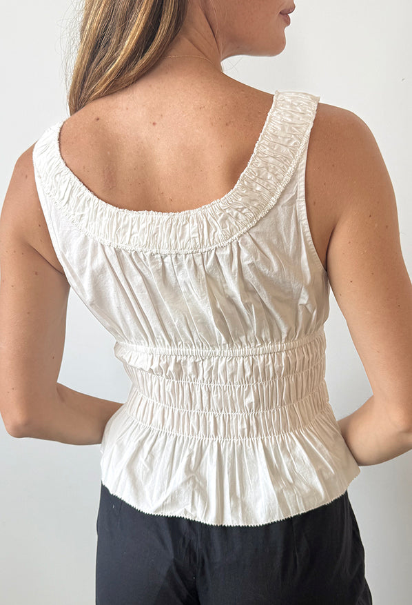 Cropped nose to waist of woman wearing a white thick strapped tank top with a shirred scoop neckline and tiered rows of smocking around the waist against a white background. 