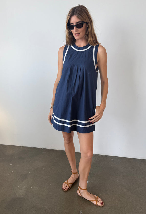 Woman in a sleeveless blue short dress with white neck, sleeve, and hem detailing standing against a plain background