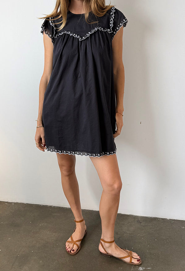 Cropped chin down of woman wearing a black mini dress with cap sleeves and white floral embroidery across the chest, sleeve and hemline standing against white background.