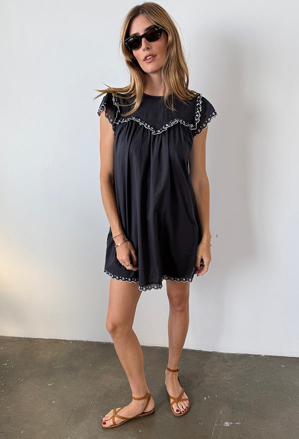 Woman wearing a black mini dress with cap sleeves and white floral embroidery across the chest, sleeve and hemline standing against white background.