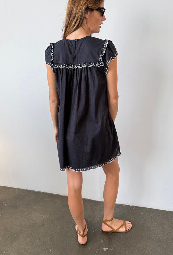 Back view of woman wearing a black mini dress with cap sleeves and white floral embroidery across the chest, sleeve and hemline standing against plain background.