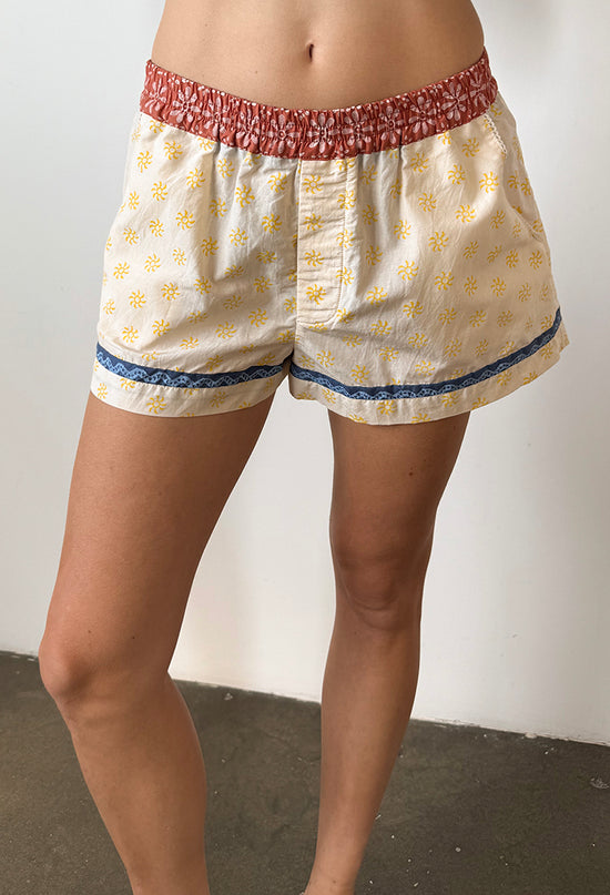 Cropped waist down of woman wearing shorts with a floral blood orange waistband and white legs with yellow sun pattern and blue hem detail on a plain background