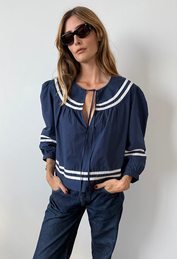 Cropped knee up woman wearing a navy blue top with white detailing paired with dark wash jeans against a plain background. 