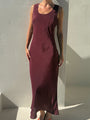 A woman modeling a sleeveless, scoop-neck maxi dress in a deep burgundy or plum shade, paired with orange sandals and black sunglasses, standing against a white stucco wall with strong sunlight and shadows.