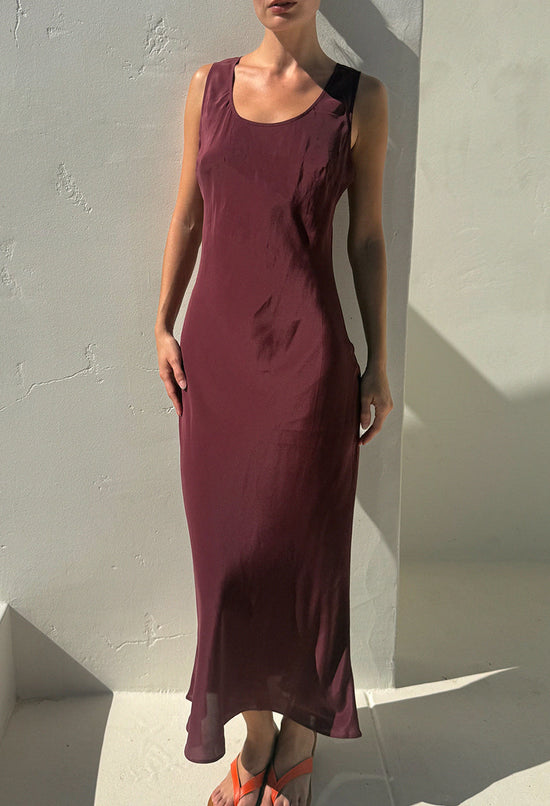 A woman modeling a sleeveless, scoop-neck maxi dress in a deep burgundy or plum shade, paired with orange sandals and black sunglasses, standing against a white stucco wall with strong sunlight and shadows.